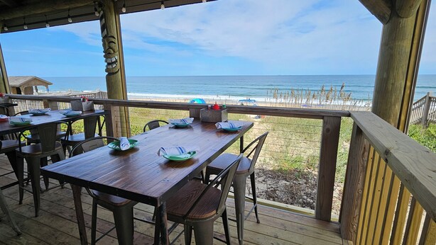Un espace de restauration en plein air, avec des tables et des chaises en bois, donnant sur une plage bordée d’une clôture en bois et offrant une vue dégagée sur l’océan.