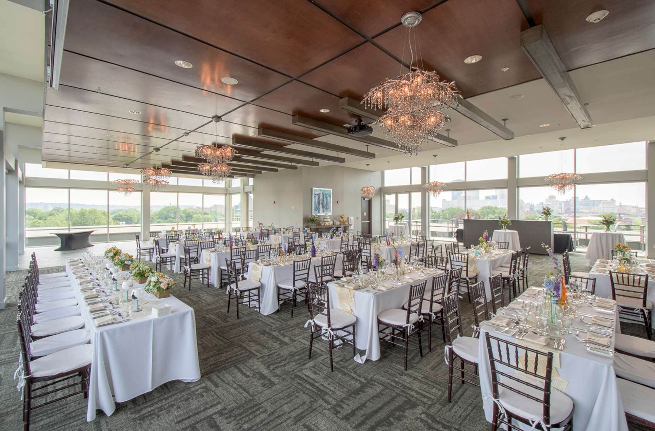 Une salle de banquet spacieuse, avec de nombreuses tables dressées pour le repas, de grandes fenêtres offrant une vue sur la ville et des lustres modernes.