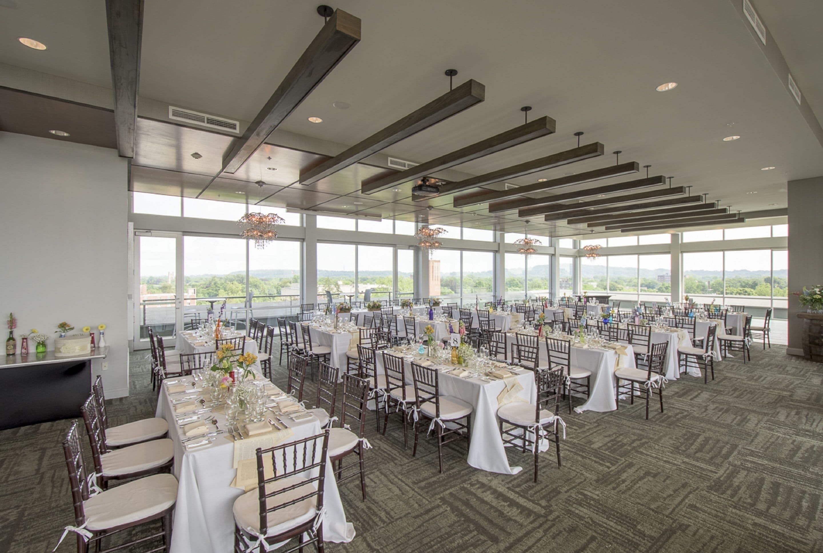A large banquet hall with multiple tables set for dining, high ceilings with exposed beams, and large windows offering a view of the outdoors.