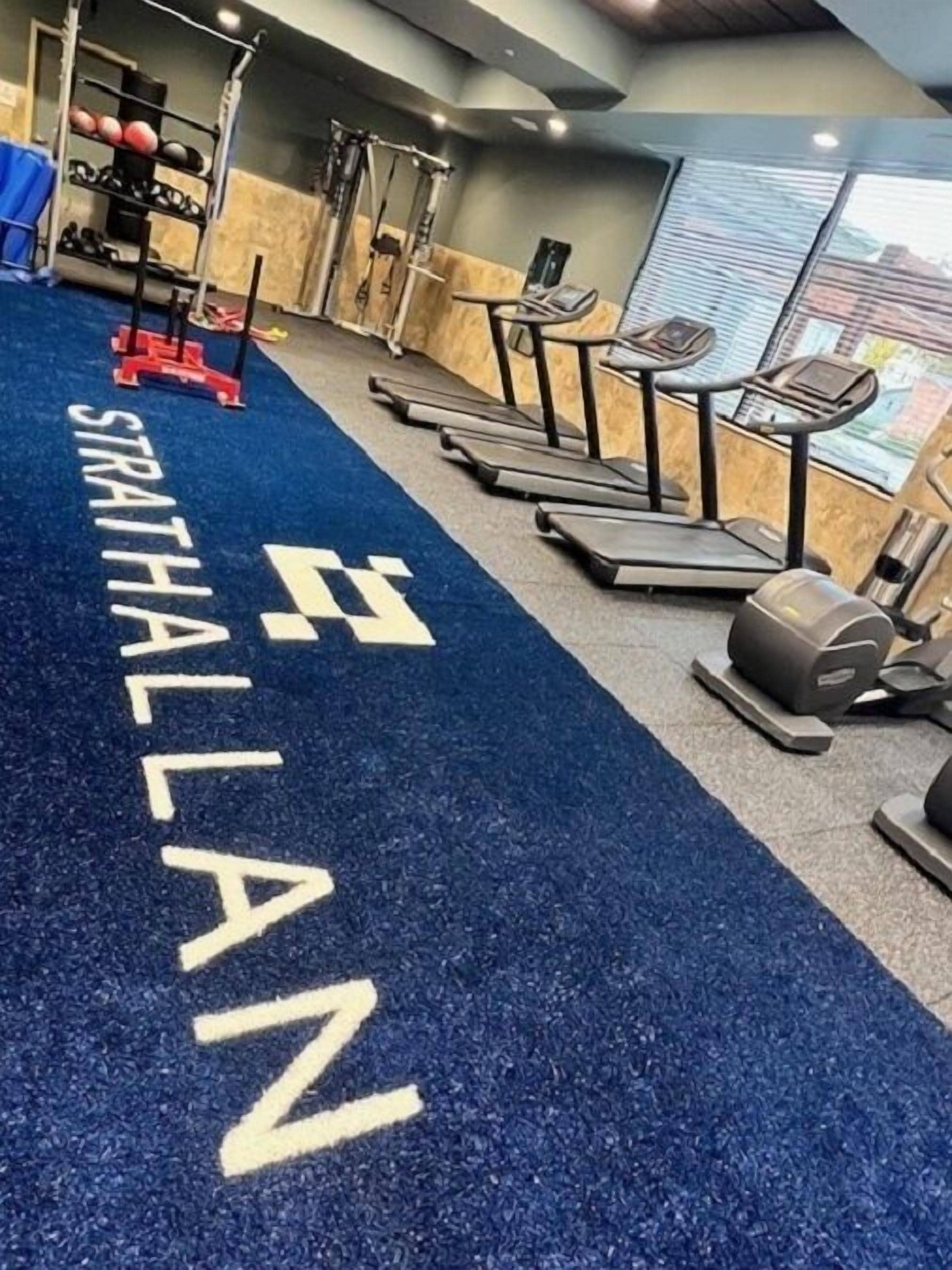 Uma academia com esteiras, aparelhos de musculação e um tapete azul com a inscrição 'STRATHALLAN'.