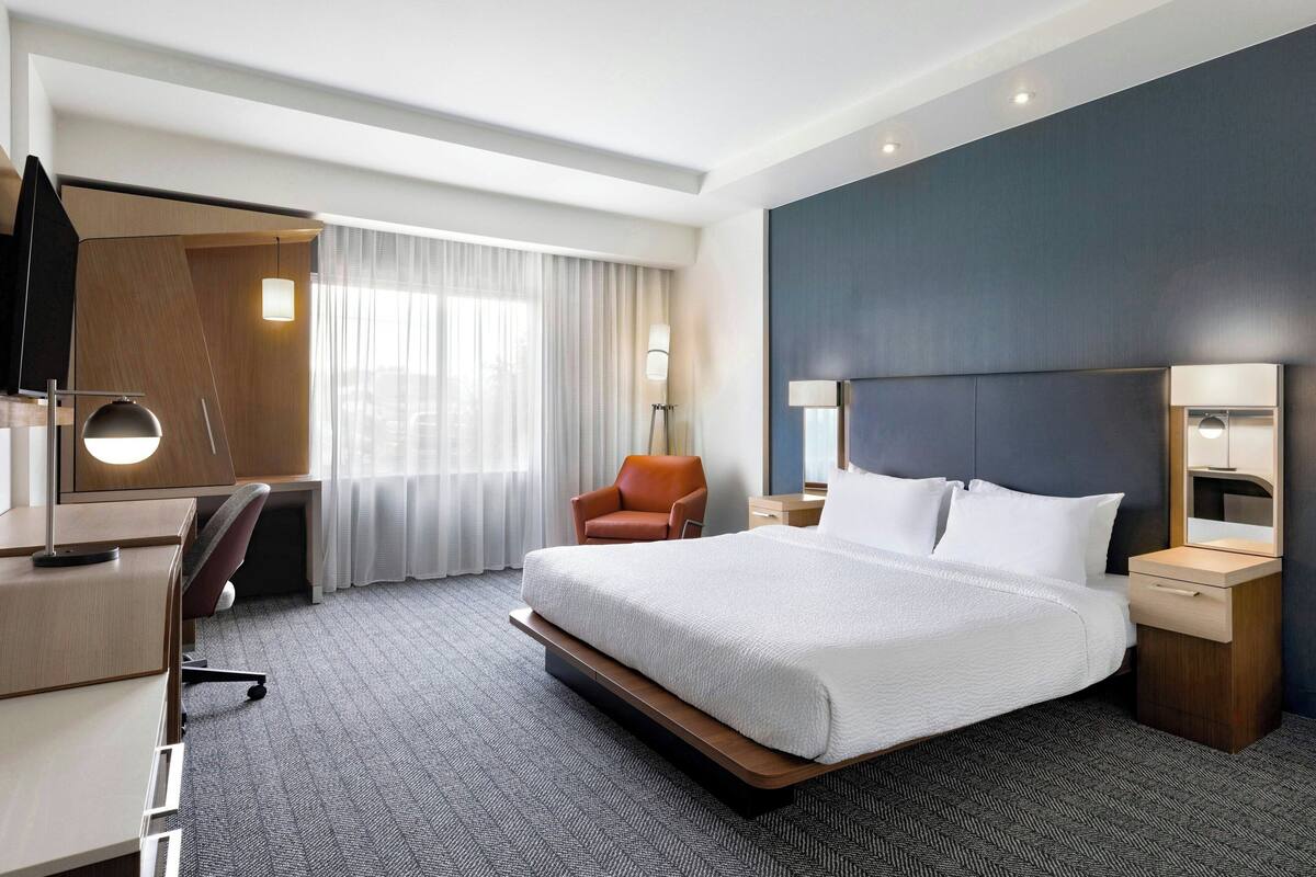 A hotel room with a large bed, a desk with a chair, a desk lamp, a chair, and a nightstand with a lamp.