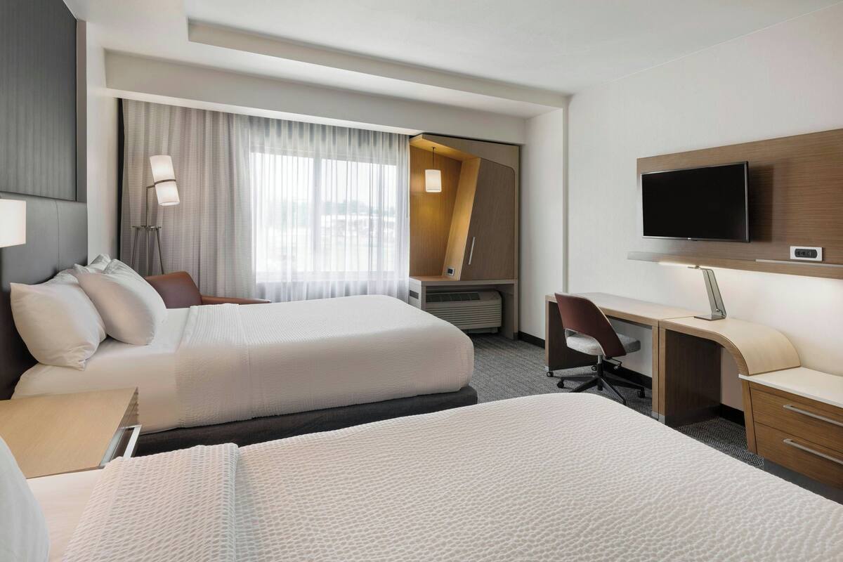 A hotel room with two beds, a desk, a chair, a TV, and a large window with curtains.