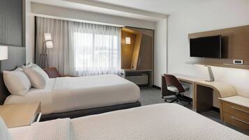 Habitación de hotel con dos camas, un escritorio, una silla, un televisor y un ventanal grande con cortinas.