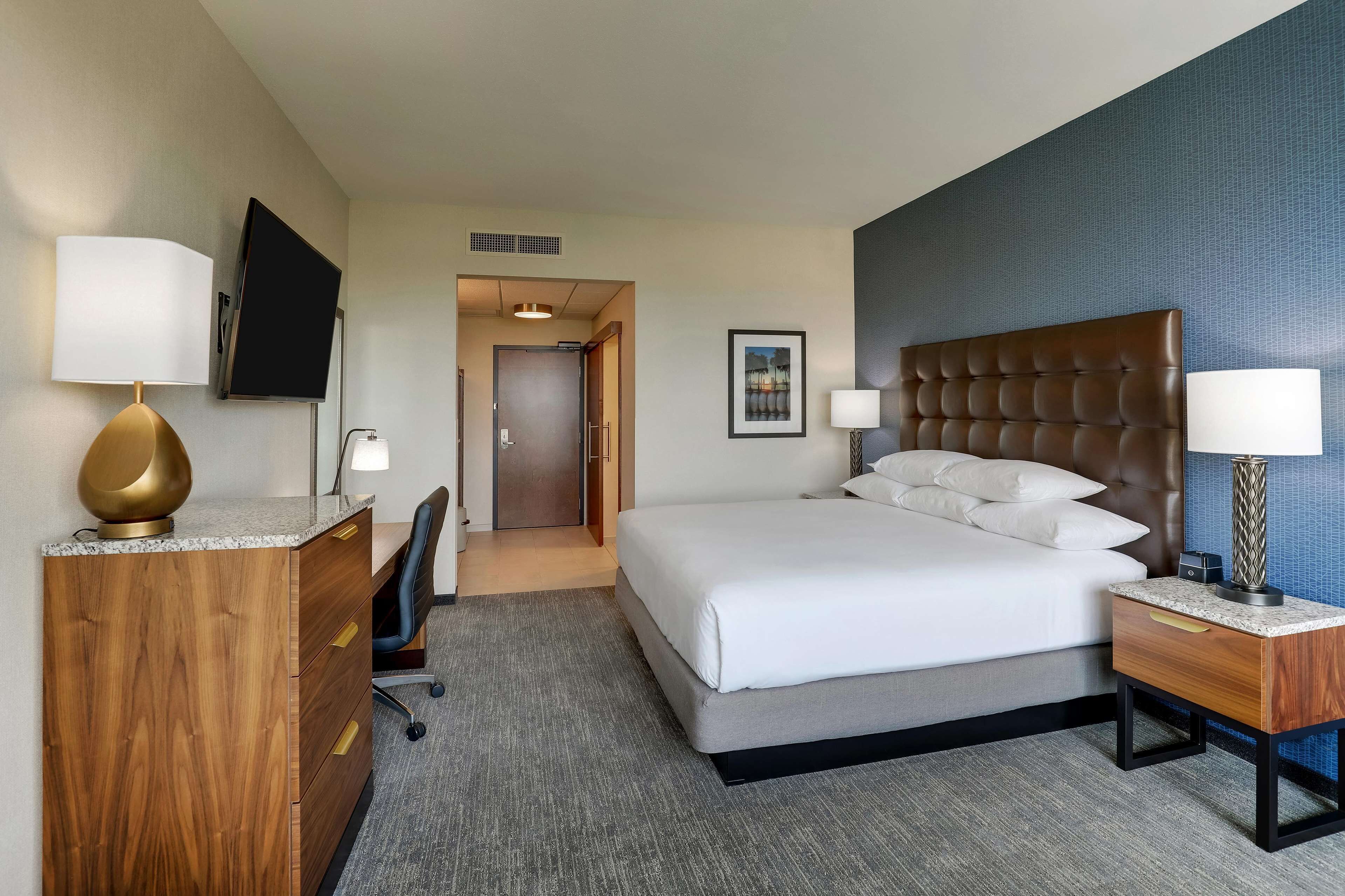 A hotel room with a bed, a desk with a lamp, a chair, and a television on the wall.