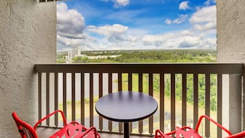 A balcony with a table and two red chairs, overlooking a green landscape and a distant building.