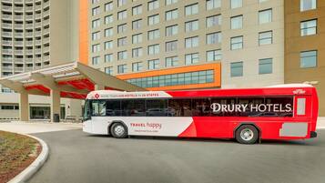 A red and white bus with "DRURY HOTELS" and "TRAVEL HAPPY" branding parked in front of a hotel building.
