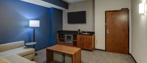 A compact hotel room with a sofa, a small table, a kitchenette, a TV, and a wooden door.