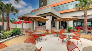 Un patio al aire libre con mesas y sillas, palmeras y un edificio con un cartel que dice "Lounge & Bar".