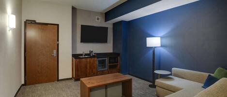 A modern living room with a sofa, a coffee table, a TV mounted on the wall, a kitchenette, and a door.