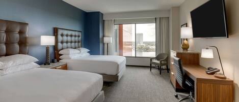 A hotel room with two beds, a desk, a chair, a TV, and a window with a city view.