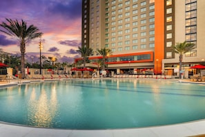 Una piscina con sillas de descanso y sombrillas, rodeada de palmeras y un edificio de hotel alto.