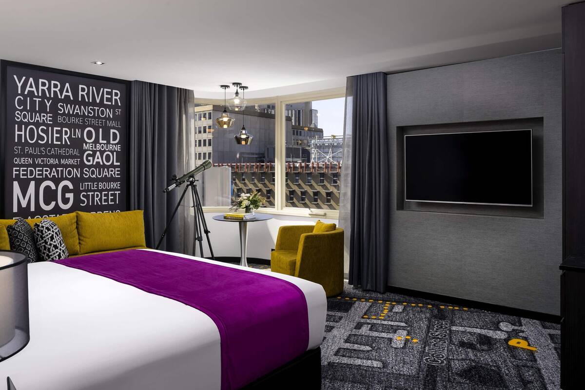 A modern hotel room with a large bed, a desk, a chair, a telescope, and a city view.