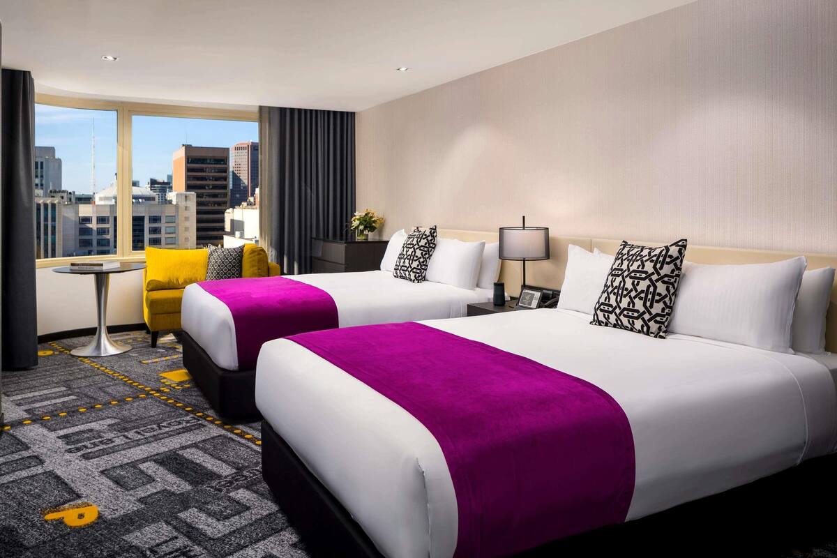 A hotel room with two beds, a small table, a chair, and a view of the city.