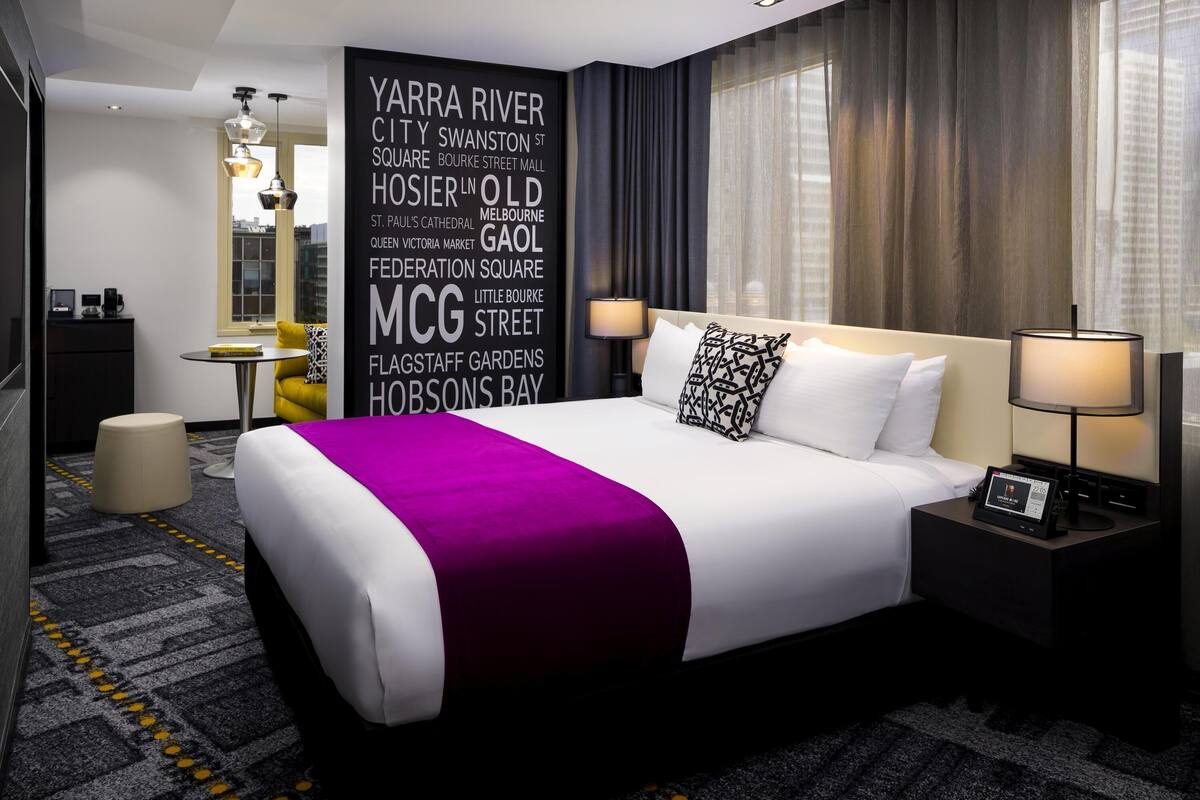 A modern hotel room with a large bed, a bedside table with a lamp, a small desk with a chair, a round table with a chair, and a wall with a cityscape and words.