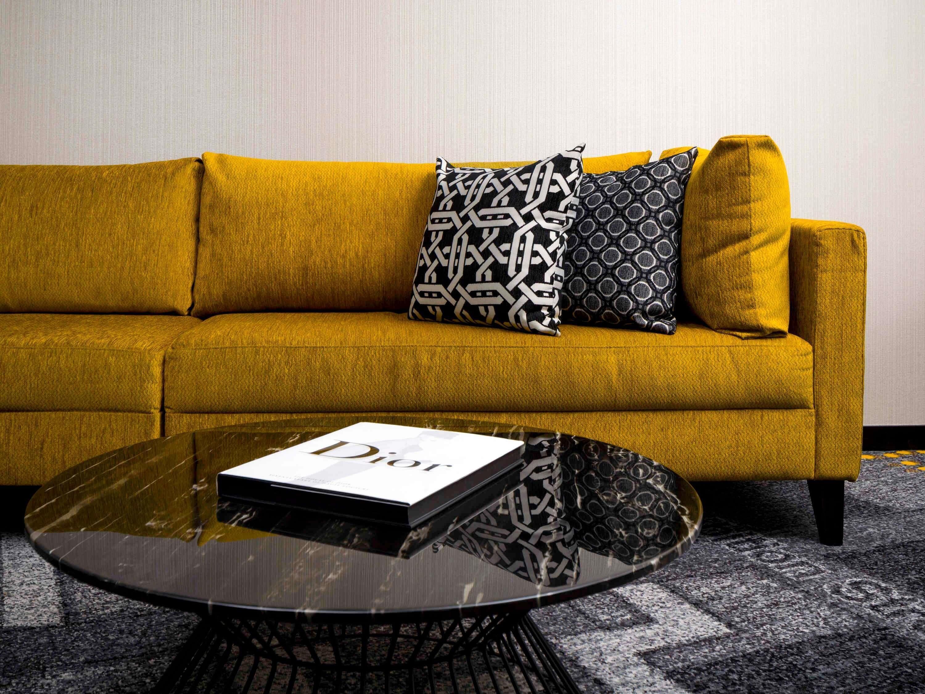 A mustard yellow sofa with patterned cushions, a glass coffee table with a book titled 'Dior,' and a carpeted floor.