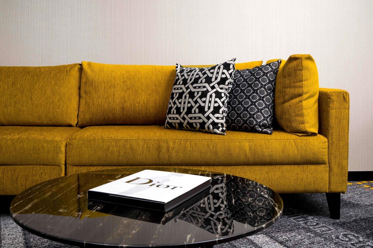 A mustard yellow sofa with patterned cushions, a glass coffee table with a book titled 'Dior,' and a carpeted floor.