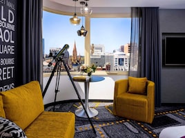 Una habitación de hotel con un telescopio, dos sillones amarillos, una mesita con un florero y un ventanal con vistas a la ciudad.