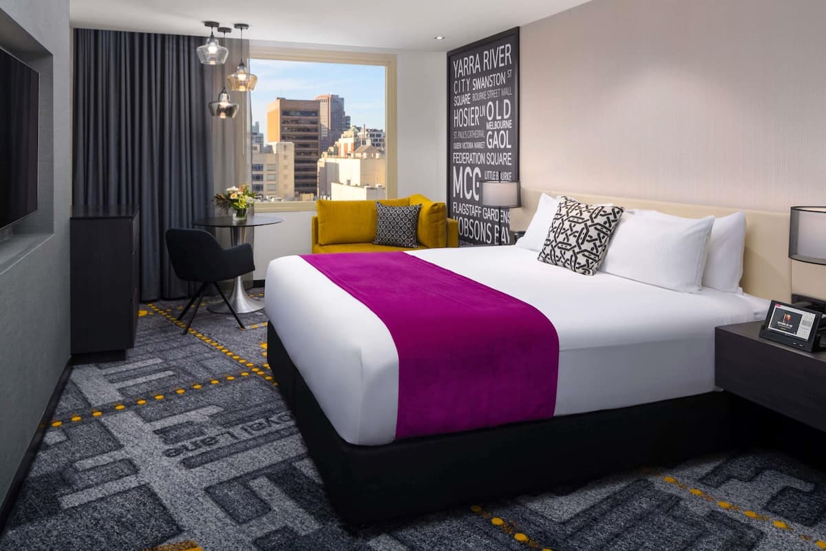 A hotel room with a large bed, a desk, a chair, a TV, and a view of the city.