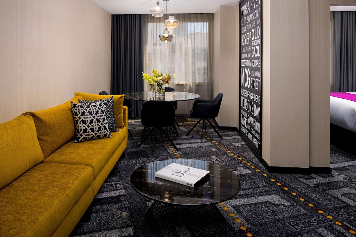 A hotel room with a mustard yellow sofa, a black coffee table, a dining area with a glass table and chairs, and a large wall with text.