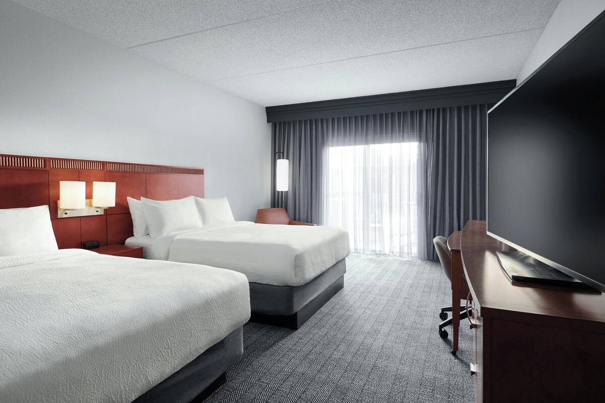 A hotel room with two beds, a desk, a flat-screen TV, and a large window with curtains.