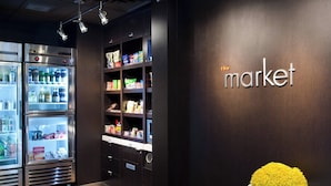A refrigerated display case with various beverages and snacks, a wall-mounted shelf with packaged goods, and a white pedestal with a yellow flower arrangement. - Courtyard by Marriott St Louis Westport Plaza (Maryland Heights)