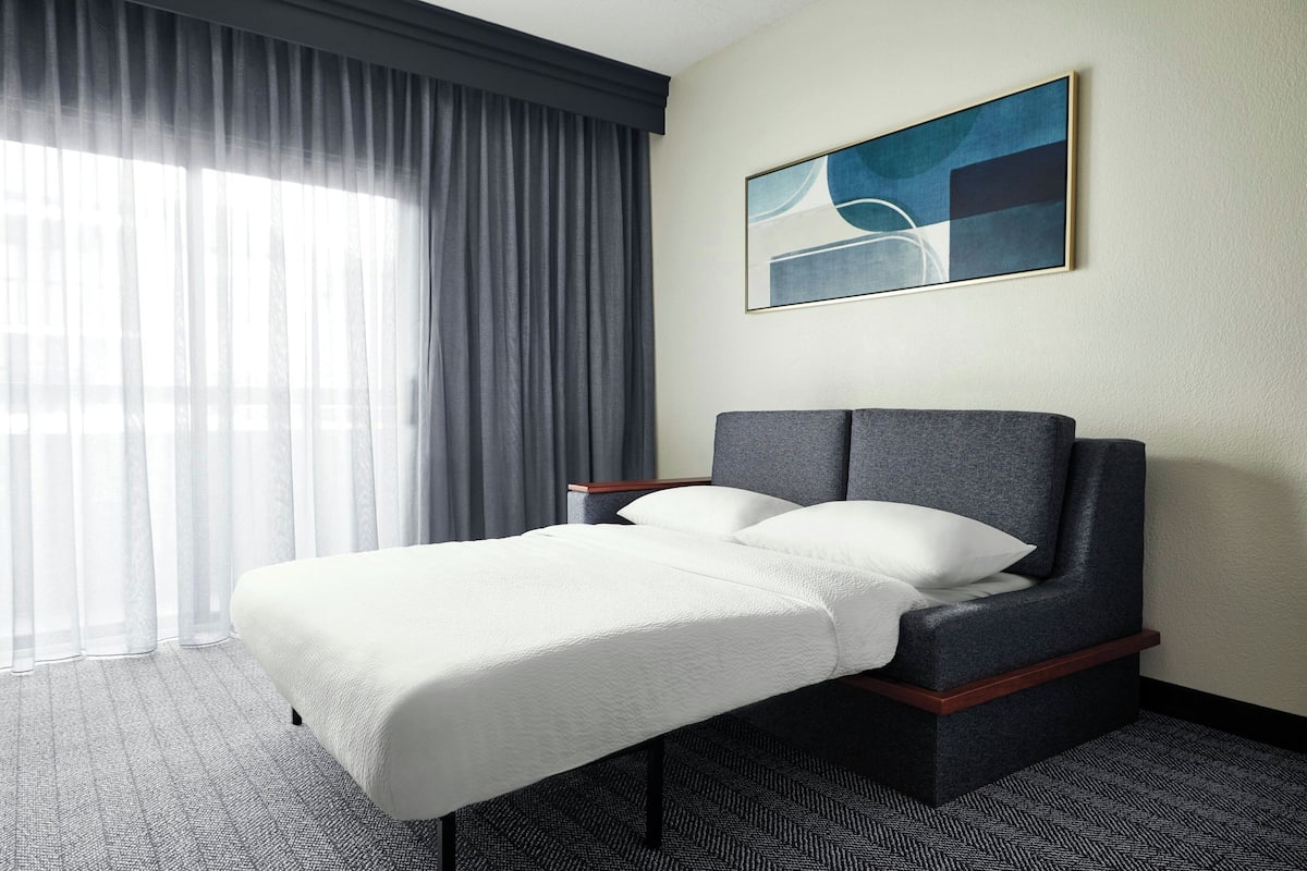A hotel room with a bed, a headboard, a window with curtains, and a painting on the wall.