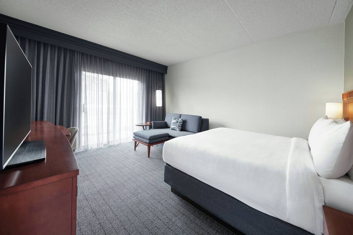A hotel room with a large bed, a desk with a flat-screen TV, a seating area with a chair and a small table, and a large window with curtains.