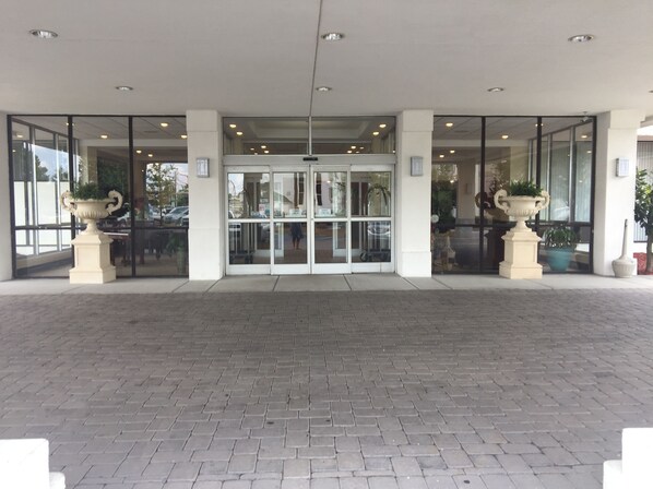 Entrada de un hotel con puertas de vidrio, plantas en macetas y un camino pavimentado.