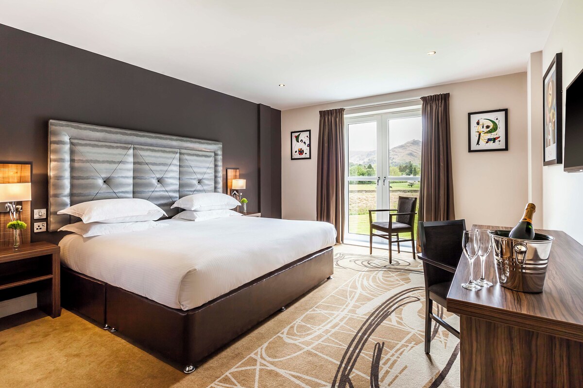 A hotel room with a large bed, a desk with a bottle of champagne, and a view of the outdoors.