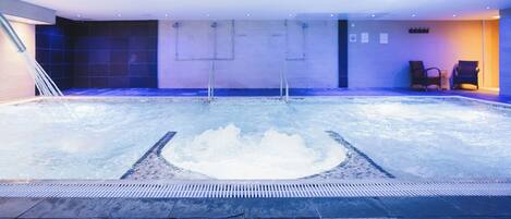 Une piscine intérieure dotée d’un grand jacuzzi central, de murs carrelés et de quelques chaises longues.