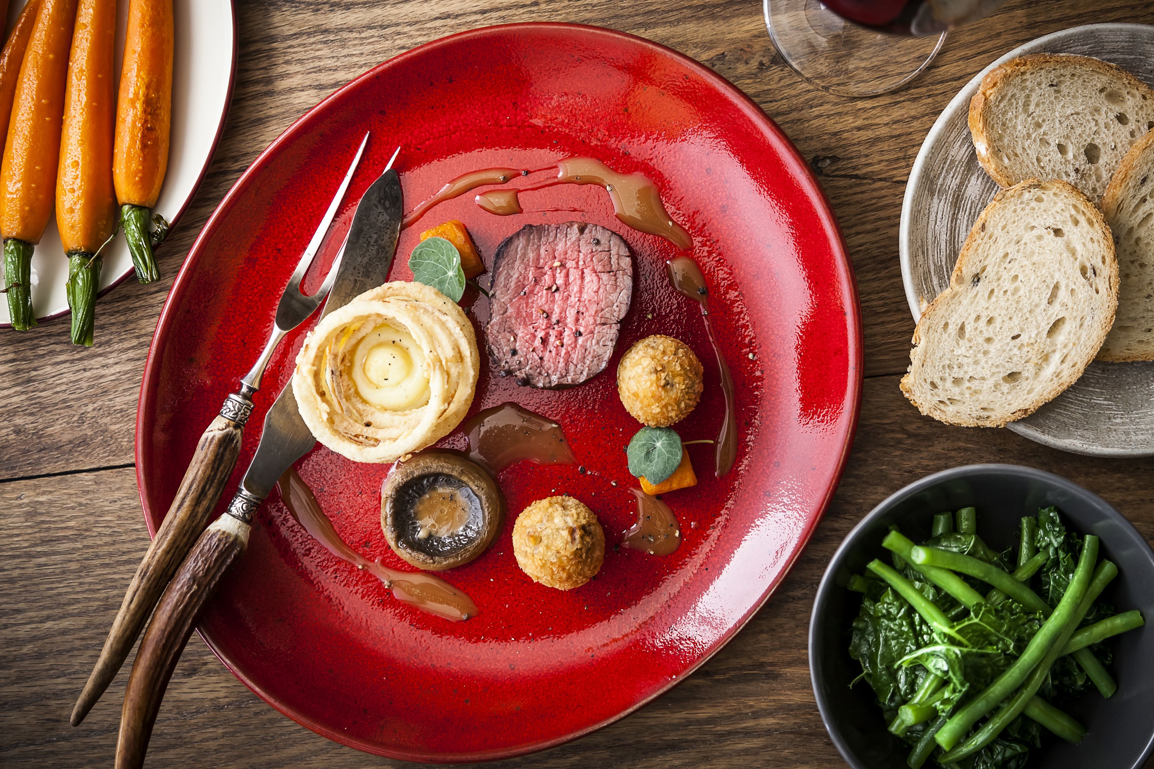 A red plate with a steak, vegetables, and bread on a wooden table.