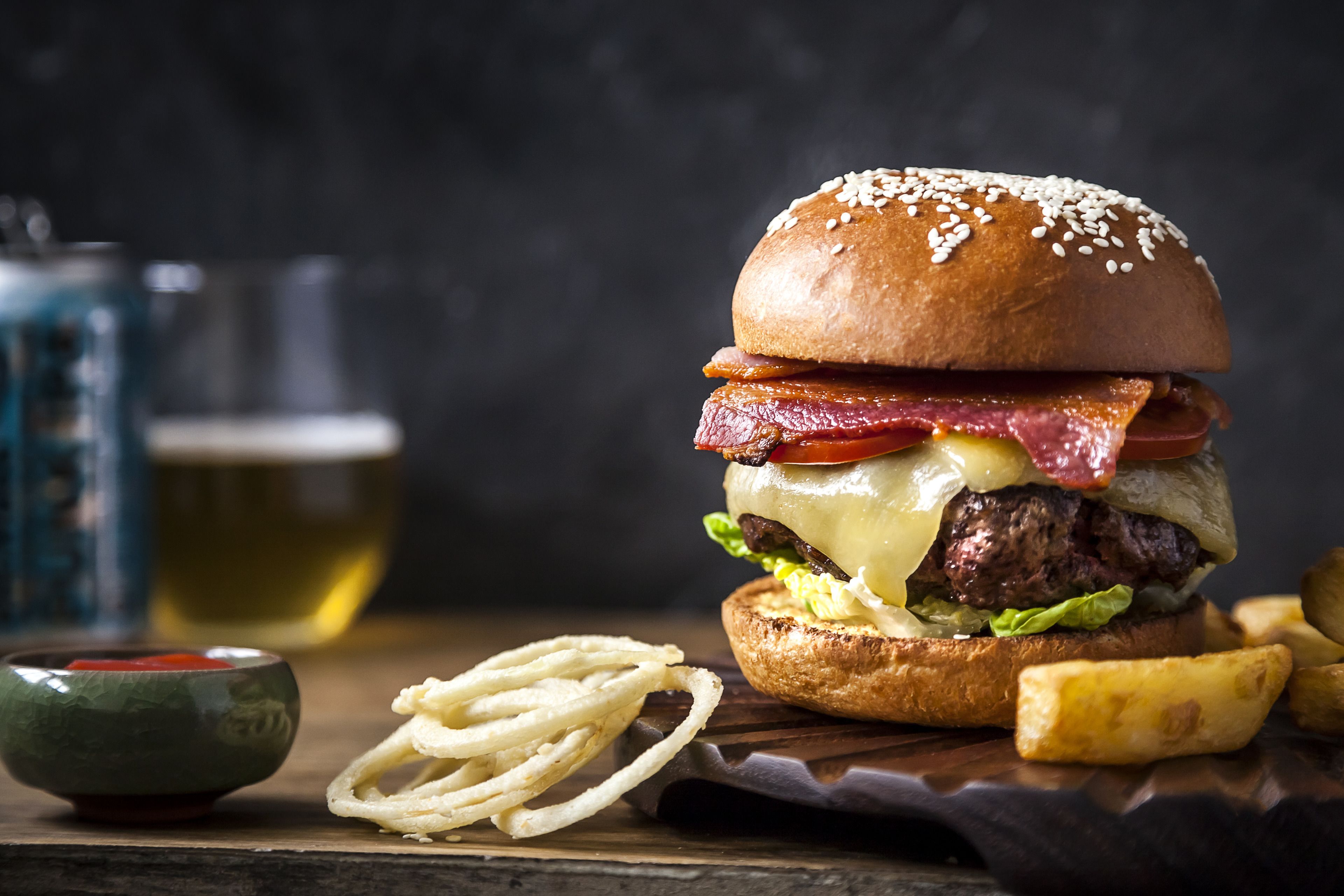 A burger with bacon, cheese, and lettuce on a bun, accompanied by fries and a small bowl of sauce.