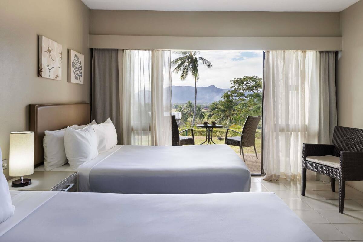 A hotel room with a large bed, a desk, a chair, and a balcony with a view of palm trees and mountains.