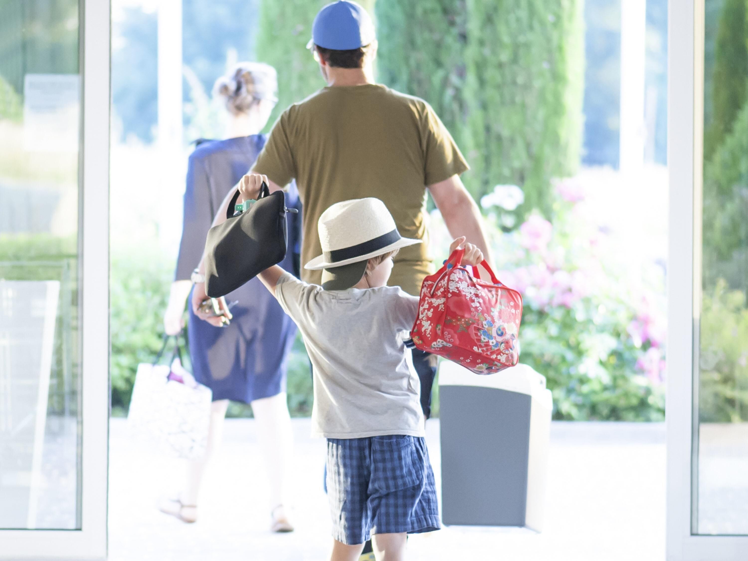 Un niño sosteniendo una bolsa roja y un sombrero, con un adulto al fondo.