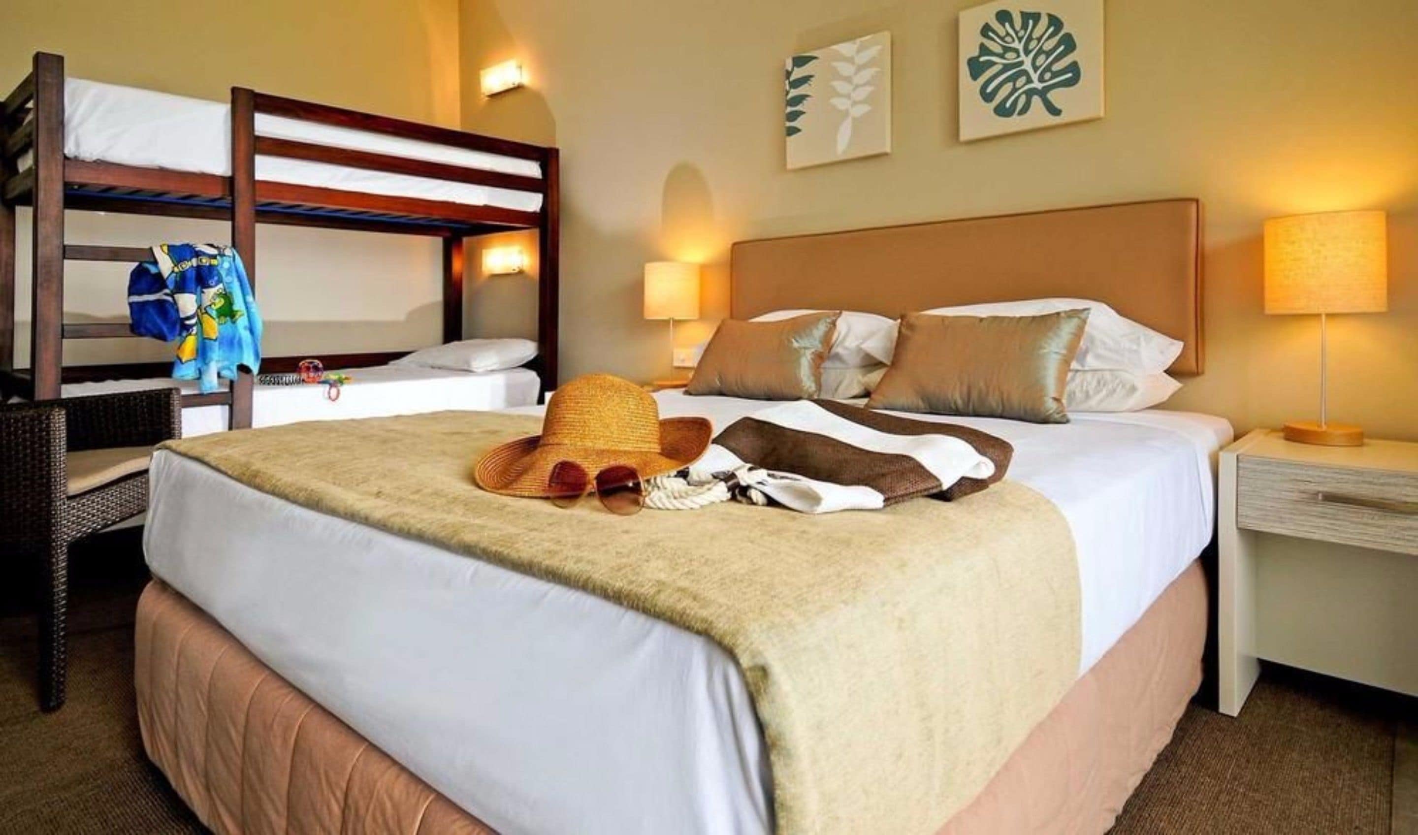 A hotel room with a bunk bed, a single bed with a beige blanket, a straw hat, and a pair of sunglasses on the bed. There are two wall-mounted lamps on either side of the bed.