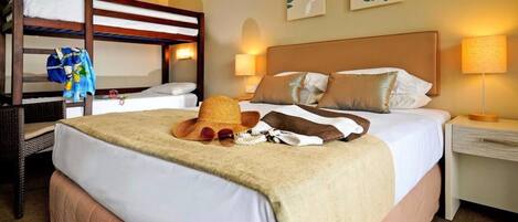 A hotel room with a bunk bed, a single bed with a beige blanket, a straw hat, and a pair of sunglasses on the bed. There are two wall-mounted lamps on either side of the bed.