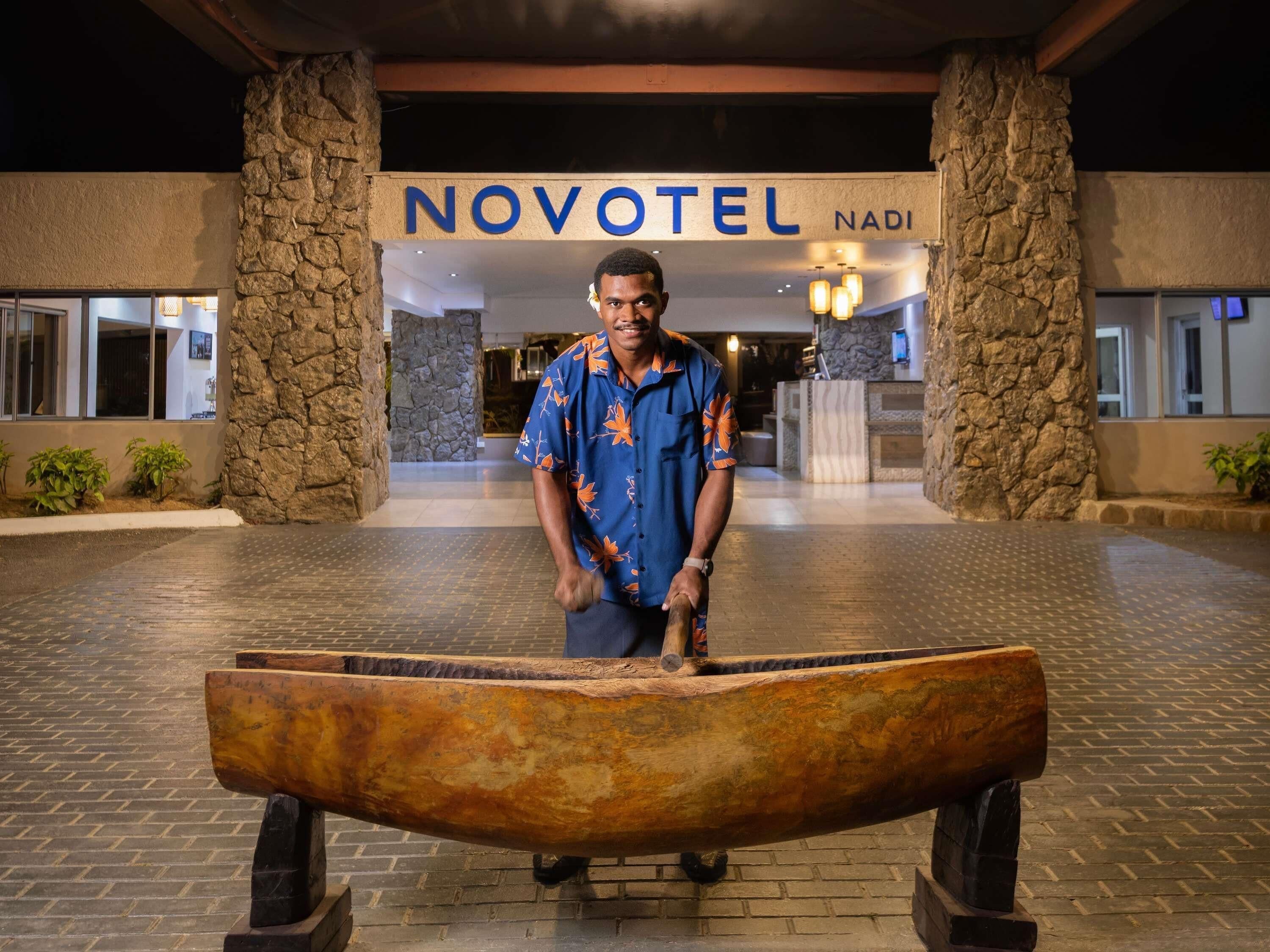 Un hombre está de pie frente a la entrada de un hotel con un cartel que dice 'NOVOTEL NADI'.