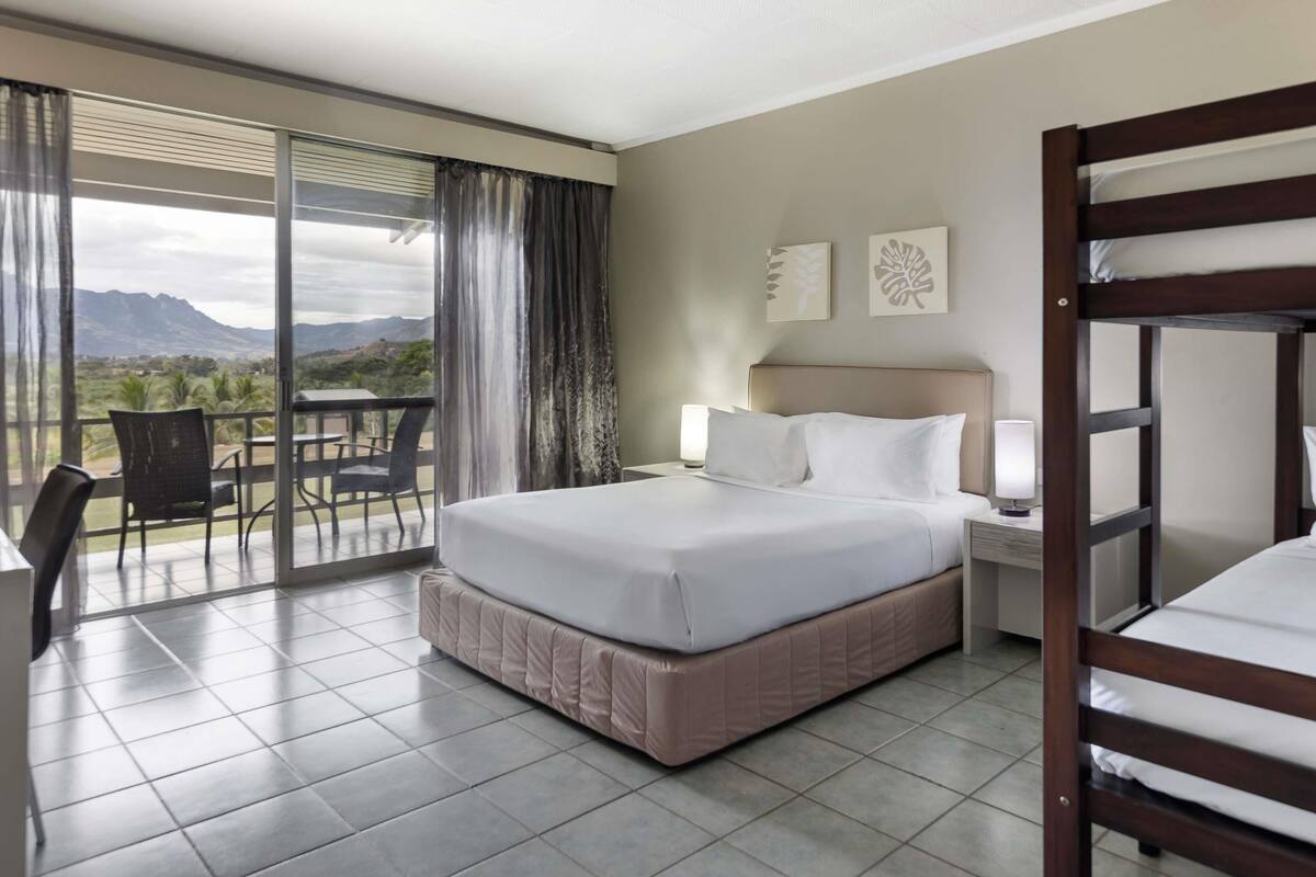 A hotel room with a large bed, a bedside table with a lamp, a bunk bed, a balcony with a table and chairs, and a view of mountains.