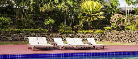 A poolside area with lounge chairs, a stone wall, and tropical vegetation.