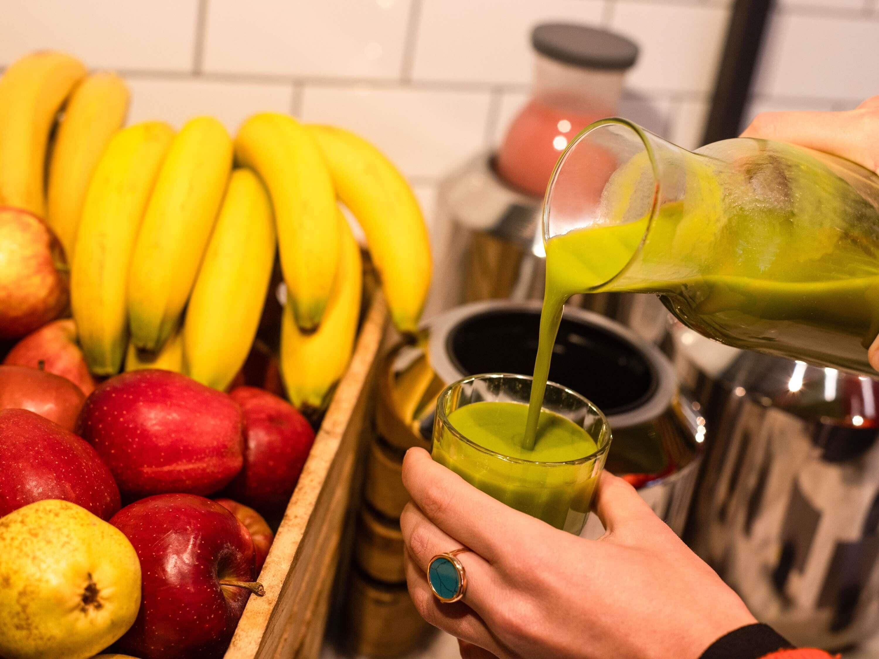 A hand pouring a green liquid into a glass, with a wooden crate of red and yellow apples and bananas in the background.