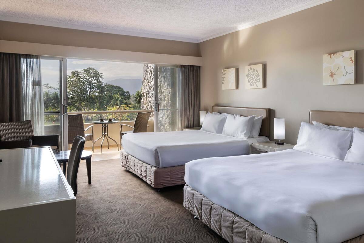 A hotel room with two beds, a desk, and a balcony with a view of the outdoors.