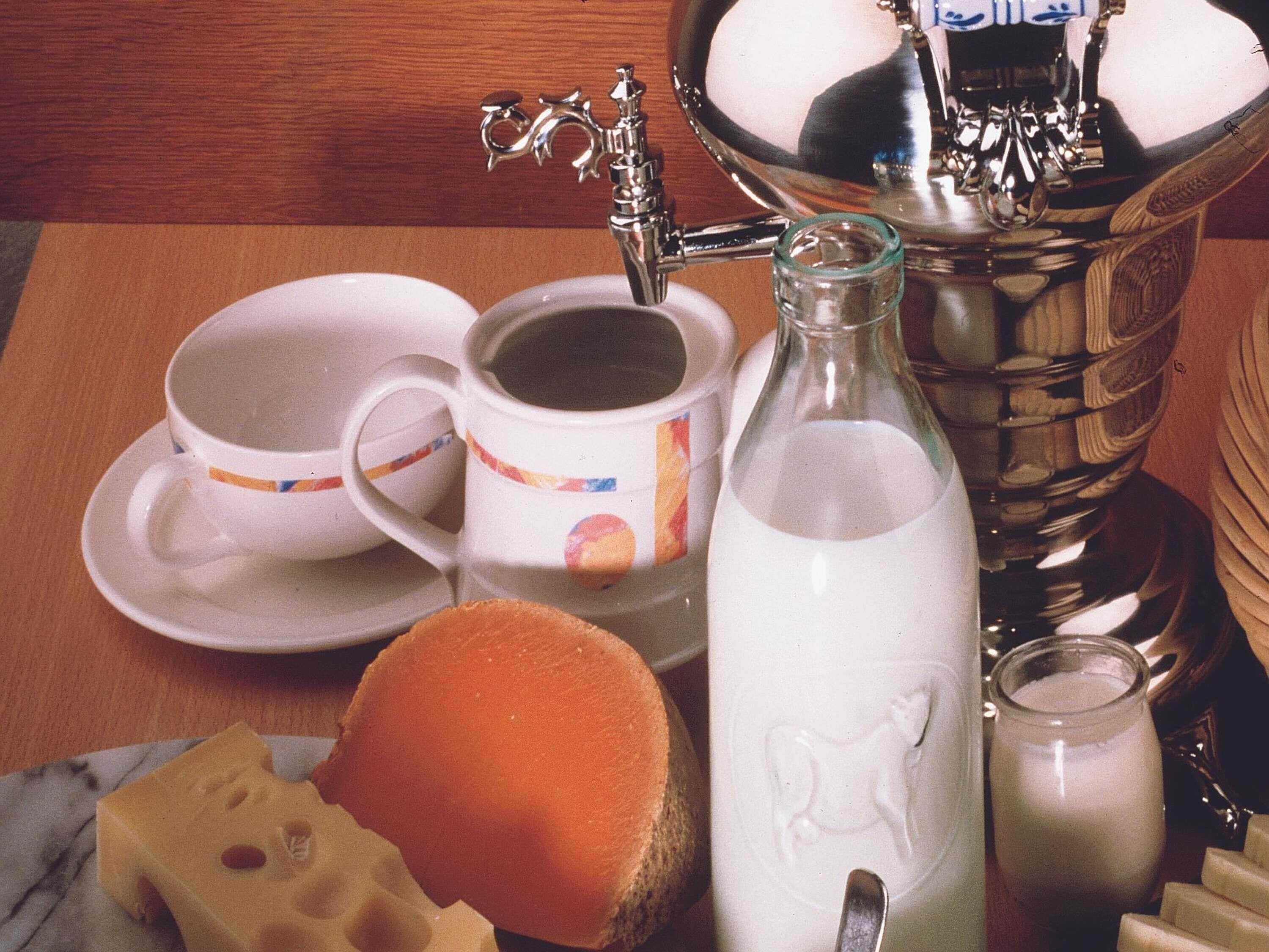 Una mesa con una cafetera, tazas, una botella, un queso redondo y un salero.