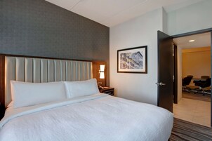 Una habitación de hotel con una cama grande, una mesita de noche con lámpara, un cuadro en la pared y una puerta abierta que da a una sala de conferencias.