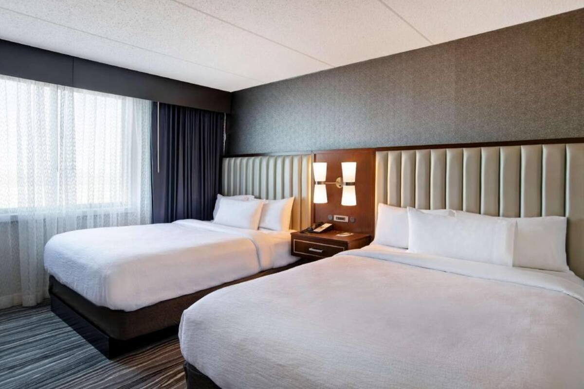 A hotel room with two beds, a headboard with a light fixture, a bedside table with a lamp, and a window with curtains.