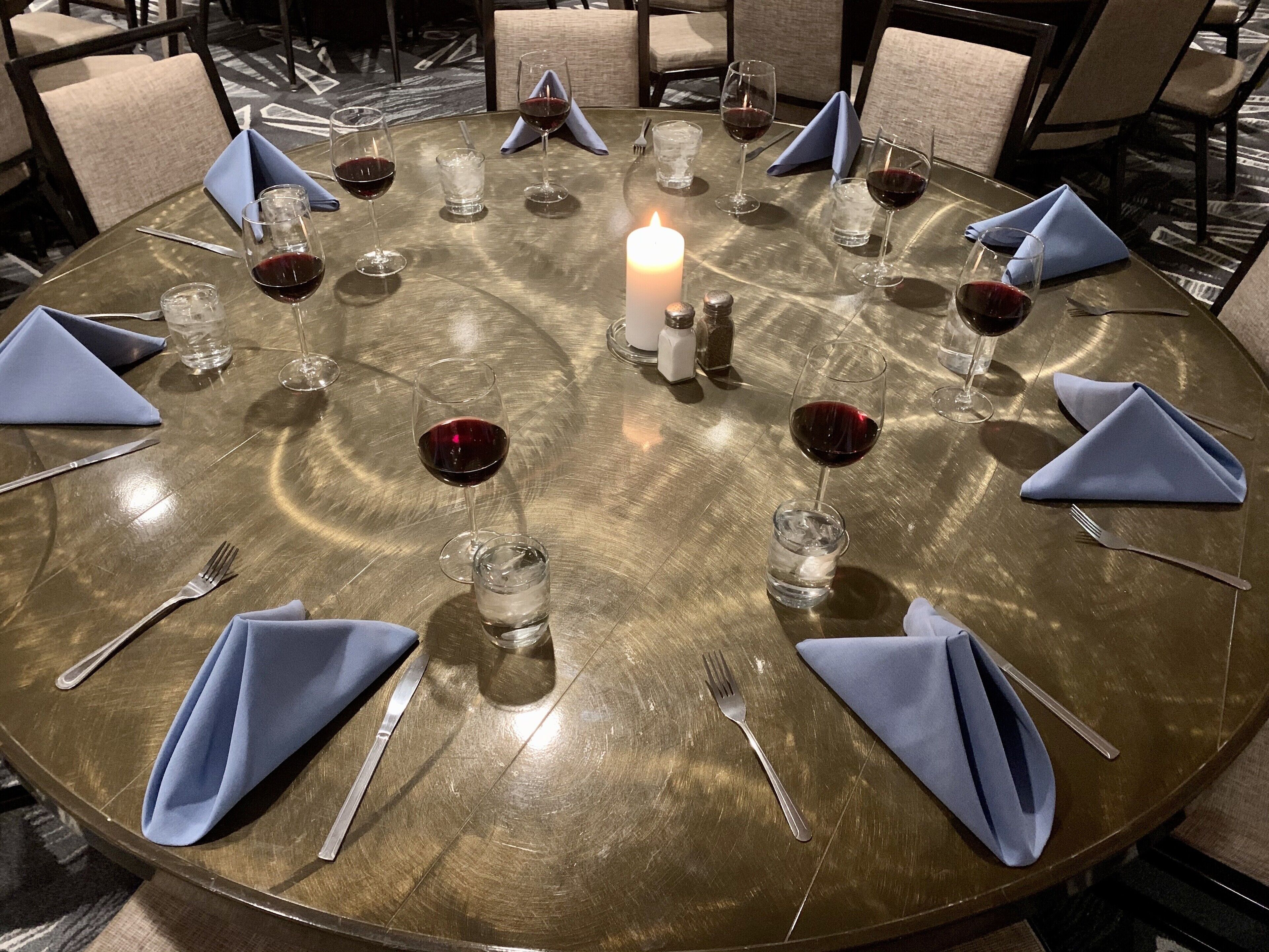 A round table with a candle centerpiece, surrounded by chairs with blue napkins and wine glasses.