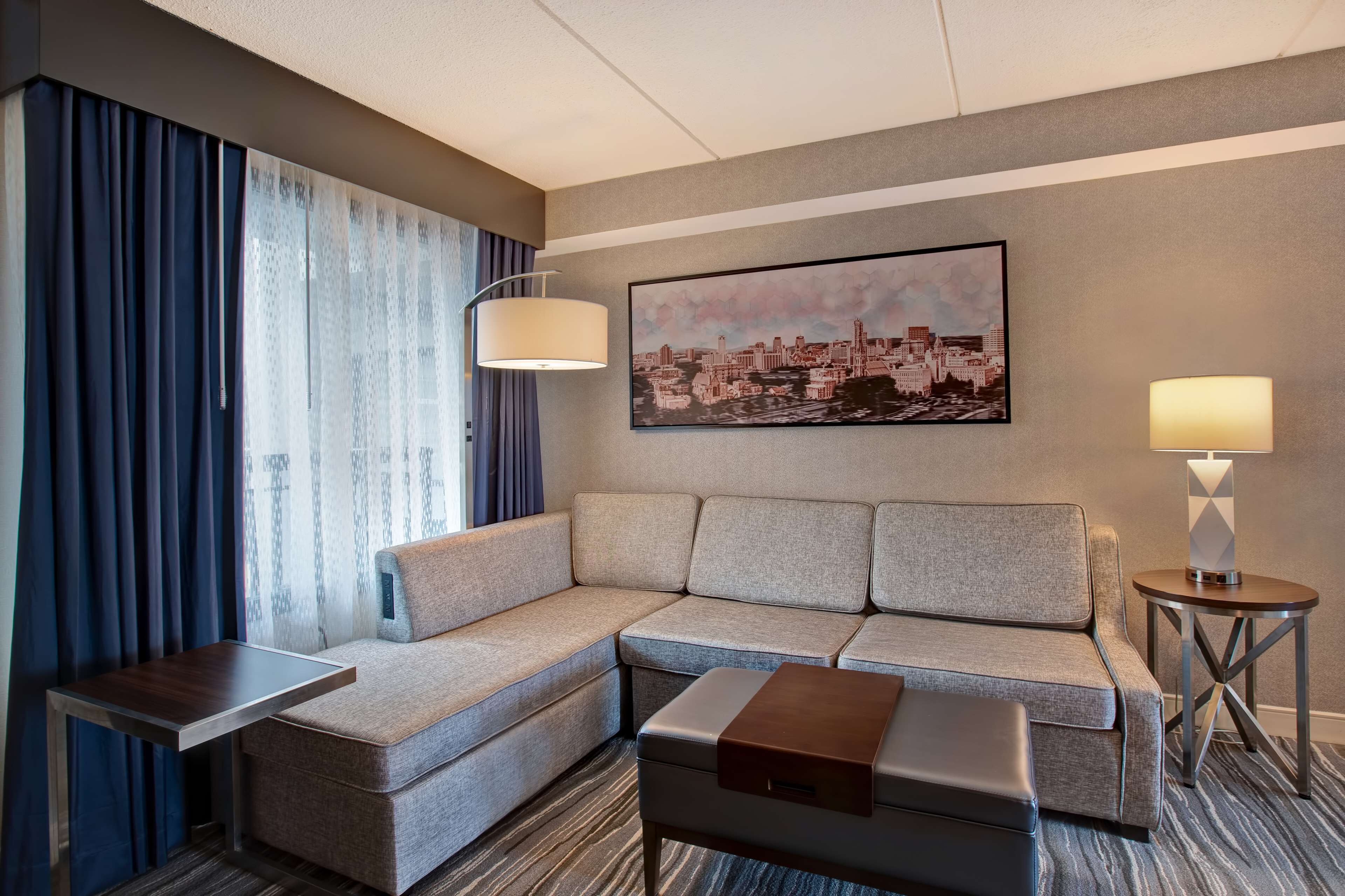 A modern hotel room with a sectional sofa, a small side table, a lamp, and a cityscape artwork on the wall.