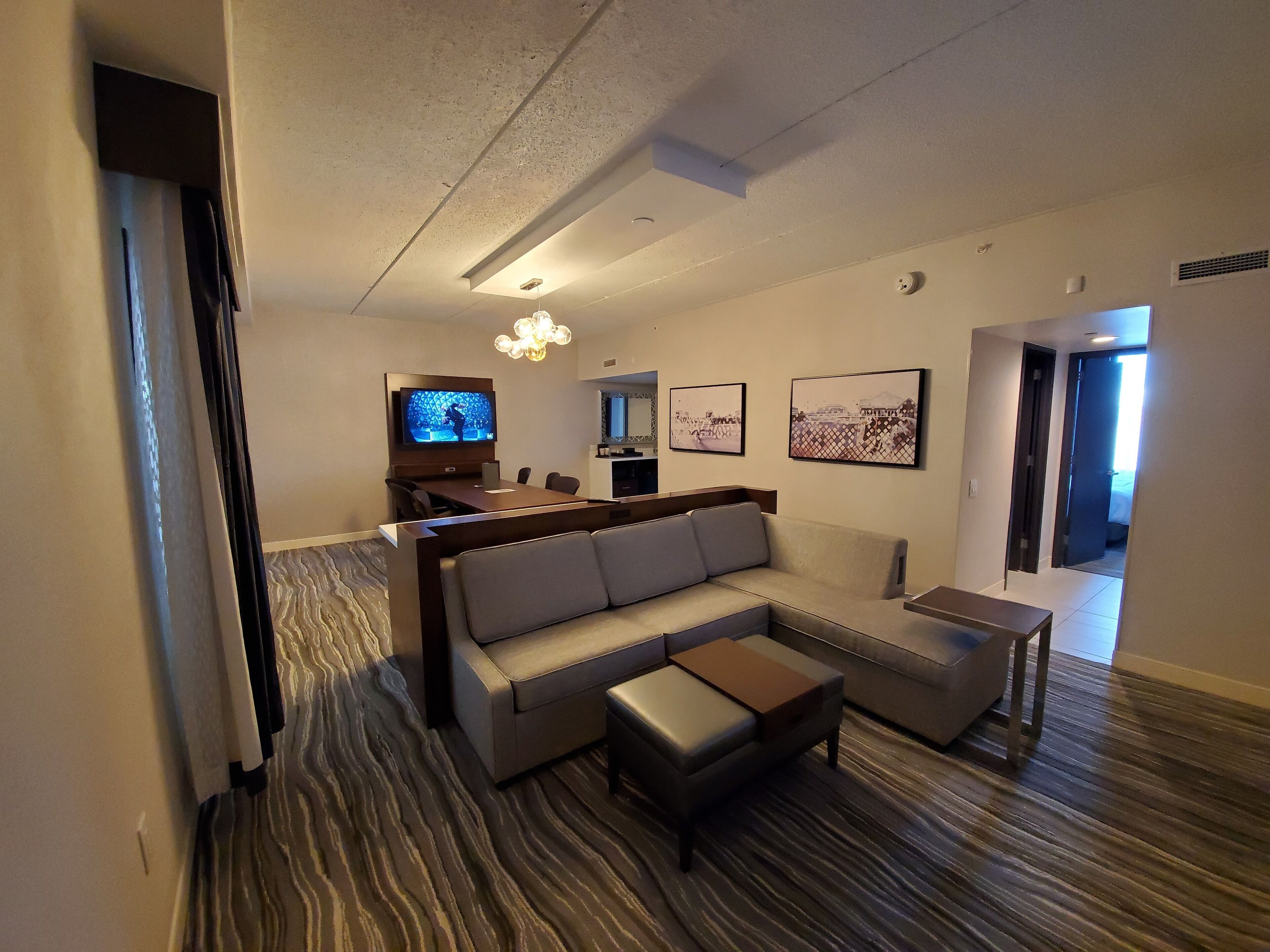 A modern hotel room with a sectional sofa, a small coffee table, a flat-screen TV, and a dining area with a bar.