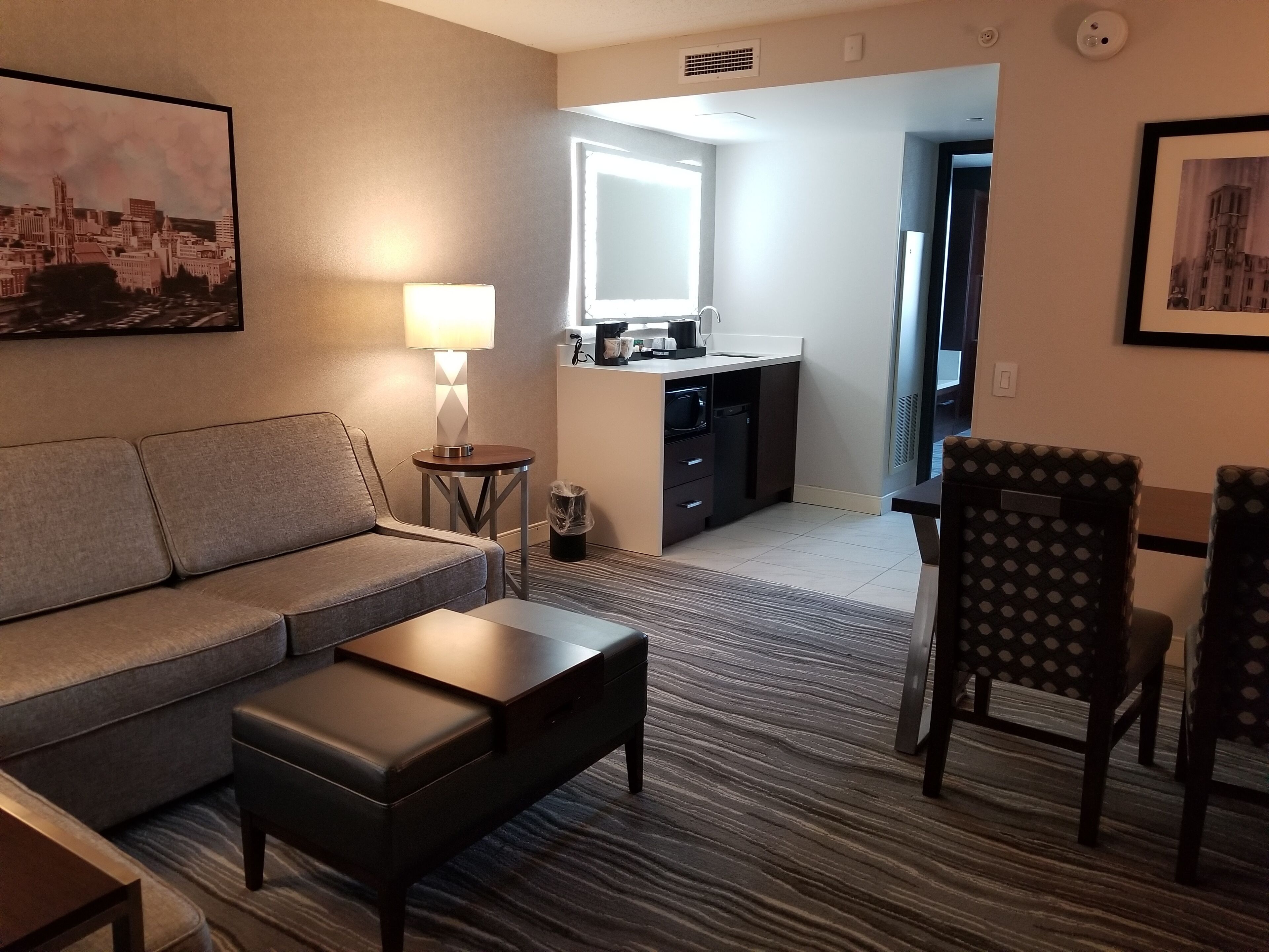 A modern hotel room with a sofa, a small table, a chair, a kitchenette, and a large window.