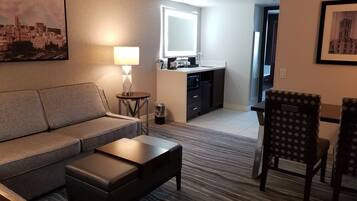 A modern hotel room with a sofa, a small table, a chair, a kitchenette, and a large window.