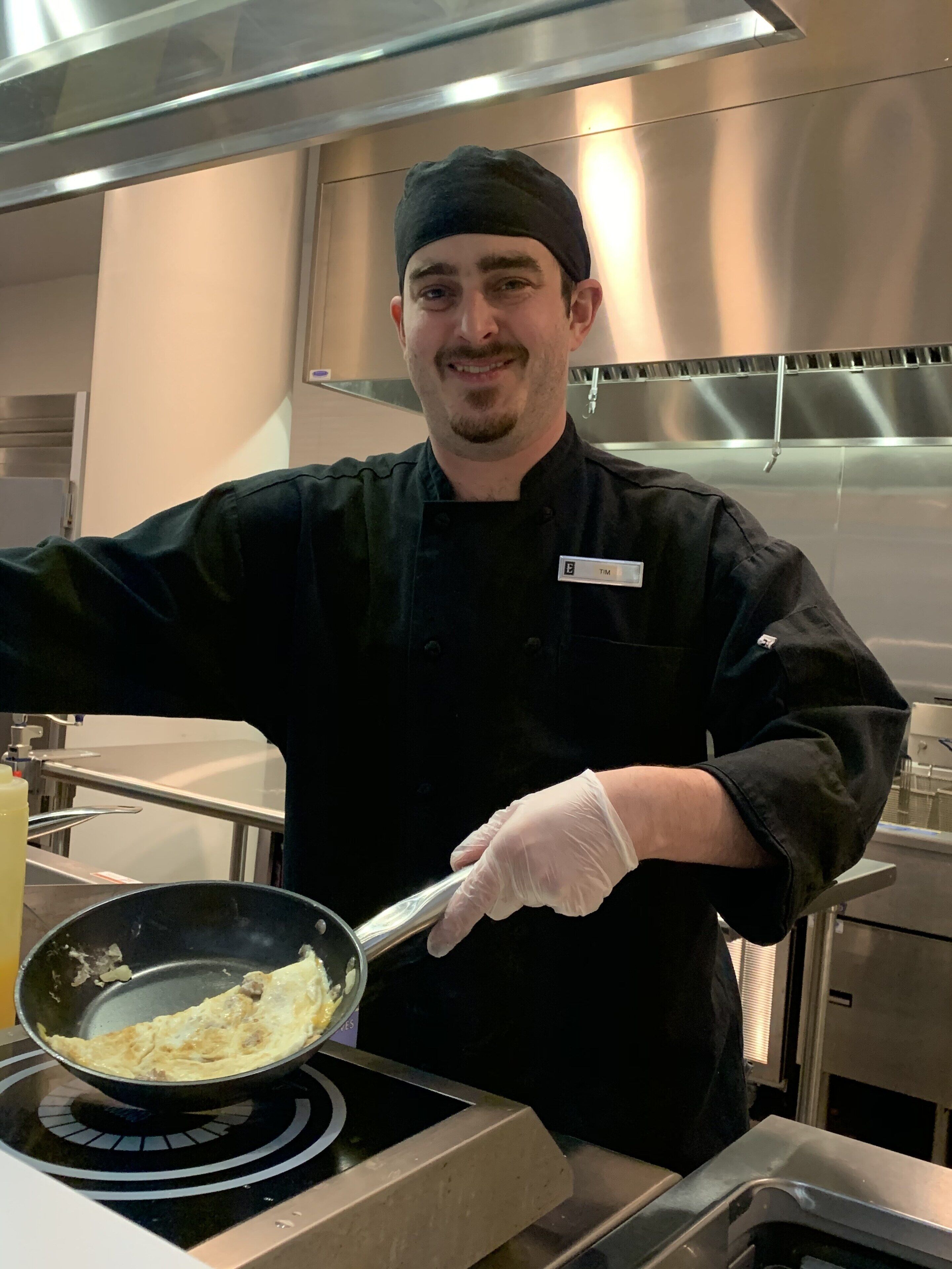 Un chef de cuisine, dans une cuisine, tenant une poêle contenant des aliments.